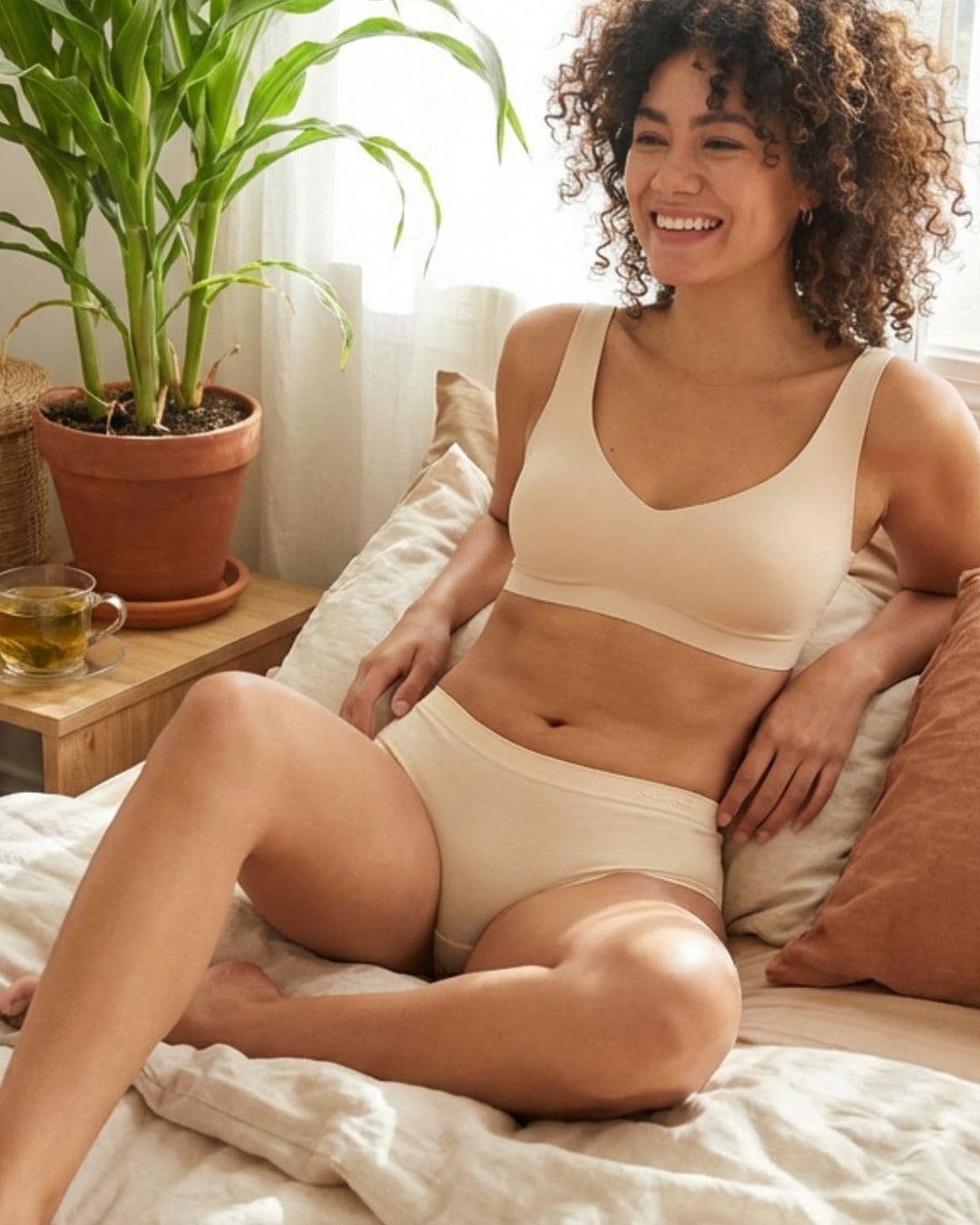 Woman wearing a beige ceramide infused wireless bra and corn bae hipster panties set sitting on a couch with a plant and tea cup in the background.