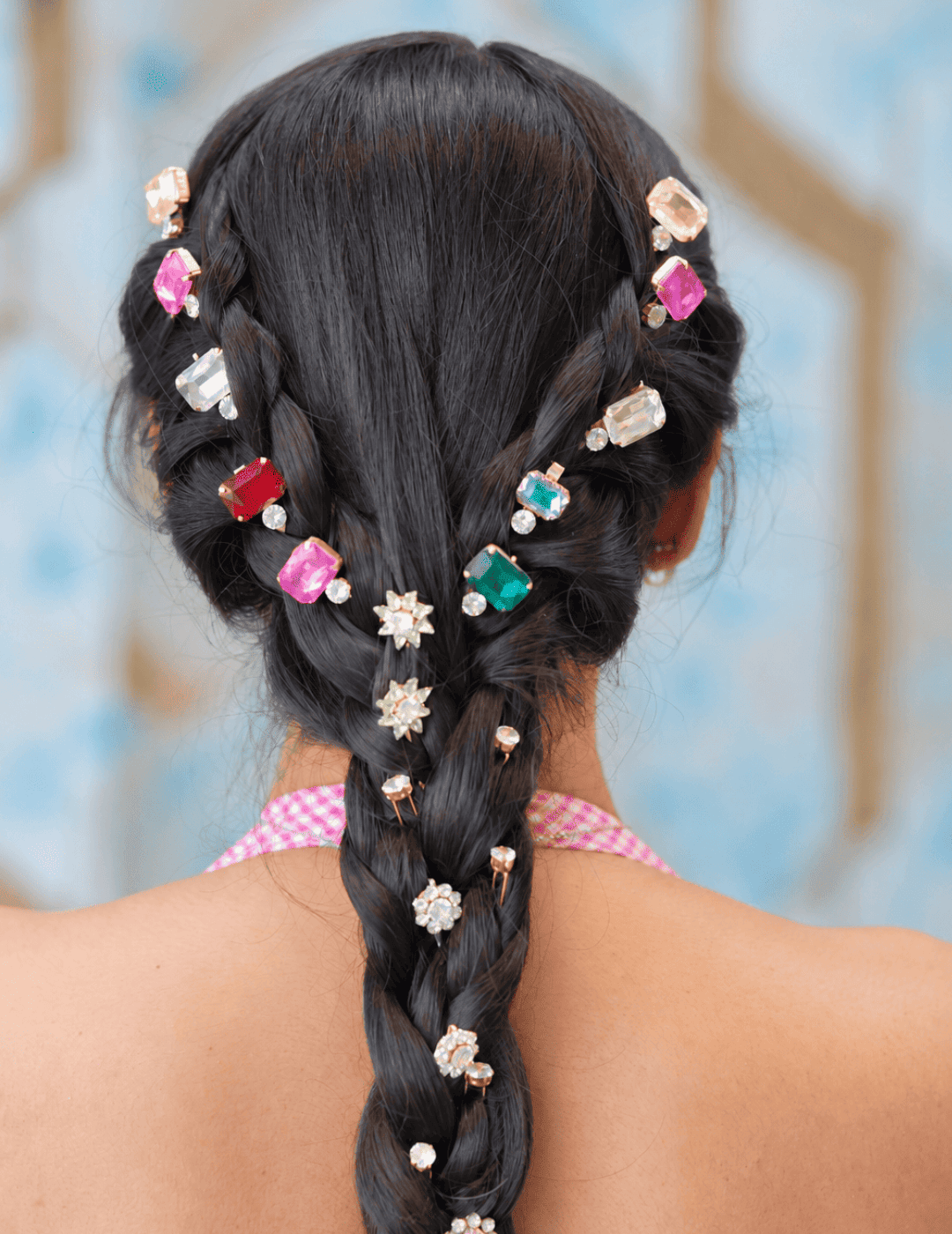A close-up back view of a person with long dark hair styled in a braided ponytail, adorned with multiple decorative hair pins featuring colorful gemstones in pink, red, green, and white, along with small white flower-shaped pins.