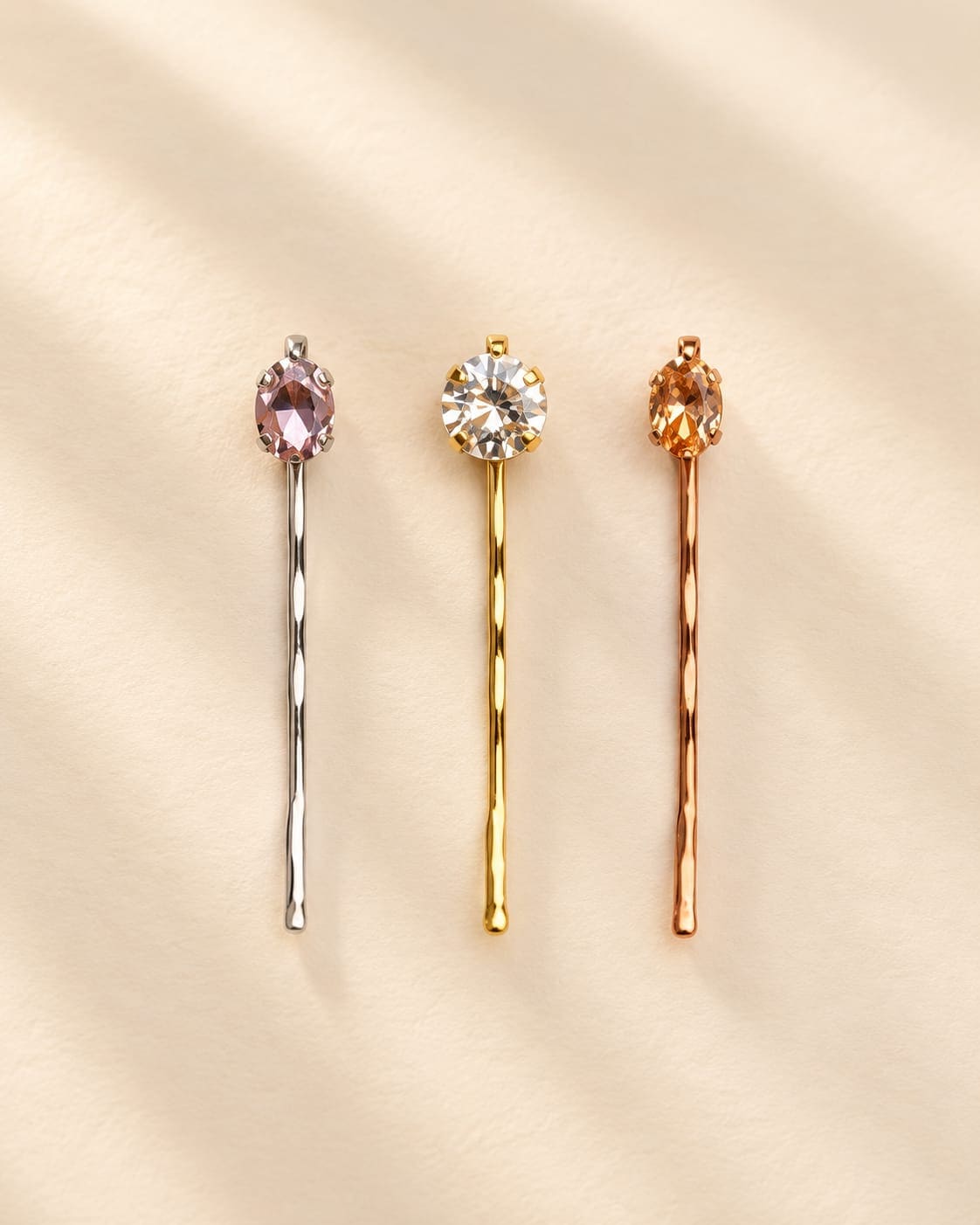 Set of 3 IQ Edit minimalist crystal bobby pins in polished silver, yellow gold and rose gold hardware finishes displaying oval pink, round clear, and oval amber gemstones against a cream background.