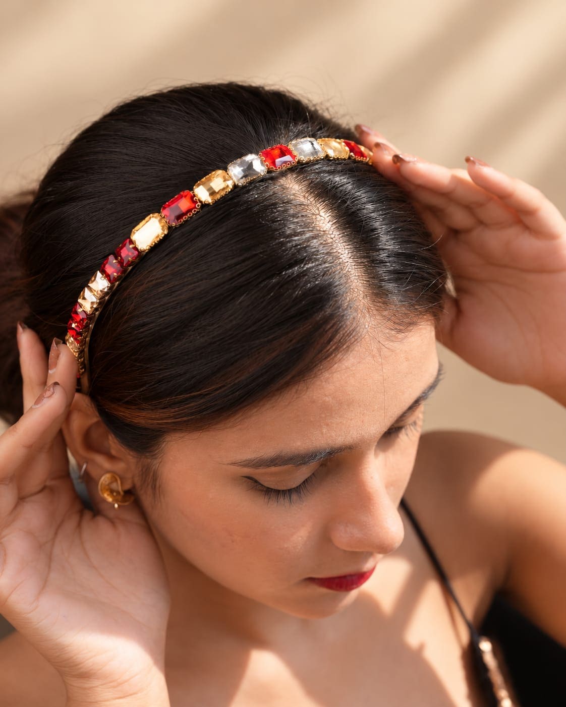 Top view of a model styling the IQ Edit Ruby Glow crystal rhinestone headband with red, clear, and champagne-toned gemstones on a sleek gold-tone band.